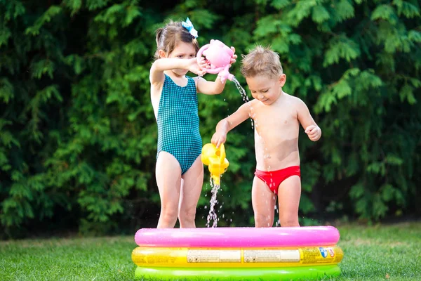 childhood summer games with water pool. Caucasian brother and sister play with plastic toys watering can pouring water splashing, inflatable round childrens bathroom. Summer hot holidays in swimsuits
