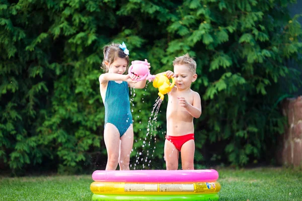Sommerspiele für Kinder mit Wasserbecken. Kaukasische Geschwister spielen mit Plastikspielzeug Gießkanne gießen Wasser spritzend, aufblasbare runde Kinderbadezimmer. Sommer heiße Ferien in Badeanzügen — Stockfoto