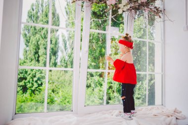 İki yaşında şık kırmızı elbiseli, kot pantolonlu ve spor ayakkabılı bir kız çocuğu pencerenin kenarındaki pencere eşiğinde duruyor. Yazın güneşli havada saç bandı ve saç stili.