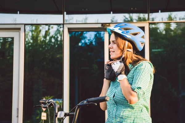 Tema da lavorare sulla moto. Una giovane donna caucasica è arrivata in bici da trasporto ecologica in ufficio. Ragazza in un edificio per uffici parcheggio biciclette in un casco, guanti e camicia e jeans — Foto Stock