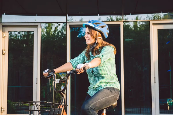 Tema da lavorare sulla moto. Una giovane donna caucasica è arrivata in bici da trasporto ecologica in ufficio. Ragazza in un edificio per uffici parcheggio biciclette in un casco, guanti e camicia e jeans — Foto Stock