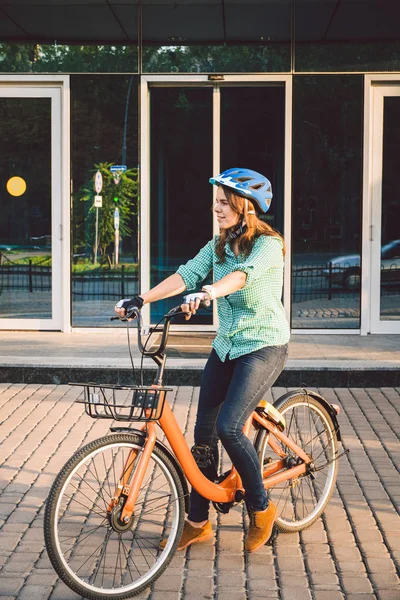 Tema da lavorare sulla moto. Una giovane donna caucasica è arrivata in bici da trasporto ecologica in ufficio. Ragazza in un edificio per uffici parcheggio biciclette in un casco, guanti e camicia e jeans — Foto Stock