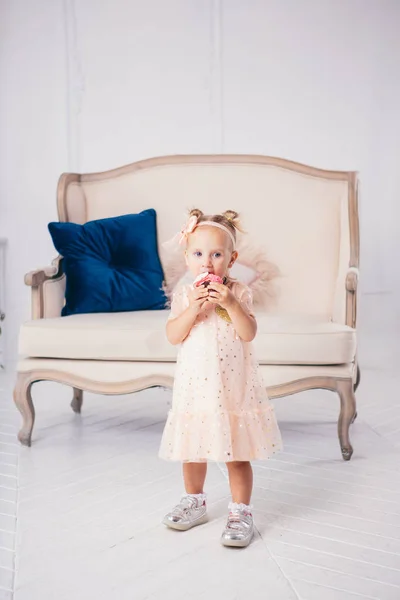 Cumpleaños de los niños. divertida chica caucásica de dos años de edad en vestido rosa de pie en el dormitorio de la casa el fondo del sofá y comer, la celebración de postre pastel. Cara y boca untadas, untadas con crema — Foto de Stock