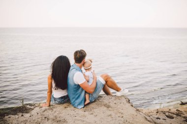 Tema doğa ve deniz küçük çocukla aile tatili. Anne, baba ve kızı bir yılın kucaklama, kızlar sırtını denize bakan sarp kayalığın yükseklikte olan adam elinde oturuyorsun