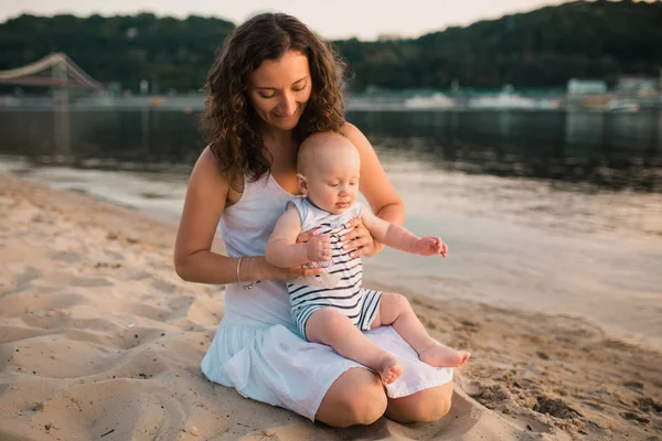 Junge Mutter sitzt mit einjährigem Sohn am Strand. Junge umarmt, lächelt, lacht, Sommertag. glückliche Kindheit unbeschwertes Spiel auf dem offenen Sand, Flusssee, Freude, Spaß. Urlaubskonzept — Stockfoto