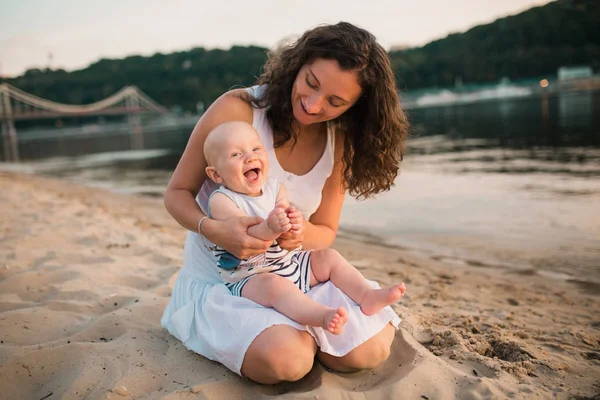 Genç anne bir yaşındaki oğluyla sahilde oturuyor. Çocuk sarılıyor, gülümsüyor, gülüyor, yaz günü. Açık kumda, nehir gölünde, neşe ve eğlence dolu çocukluk oyununuz kutlu olsun. tatil kavramı — Stok fotoğraf