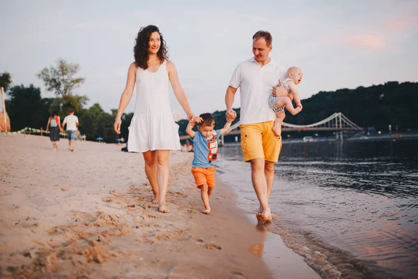 Vacaciones familiares en verano. Joven familia caucásica pie caminando descalzo playa de arena, ribera de agua del río. Papá mamá cogido de la mano dos hijos, hermanos. Gran familia amigable con dos niños cerca del lago —  Fotos de Stock