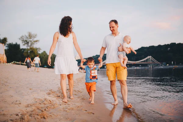 family vacation in summer. Young Caucasian family foot walking barefoot sandy beach, shore river water. Dad mom holding hands two children, brothers. Big friendly family with two children near lake