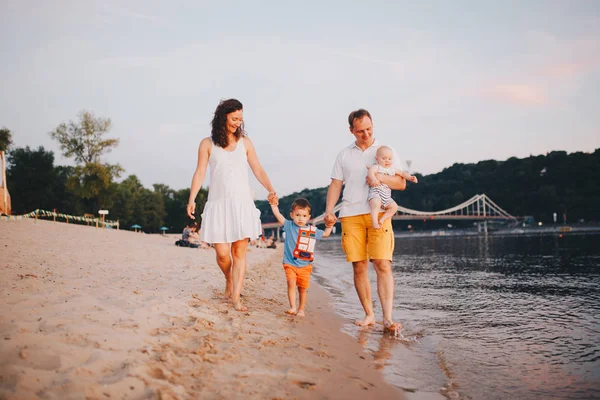 family vacation in summer. Young Caucasian family foot walking barefoot sandy beach, shore river water. Dad mom holding hands two children, brothers. Big friendly family with two children near lake