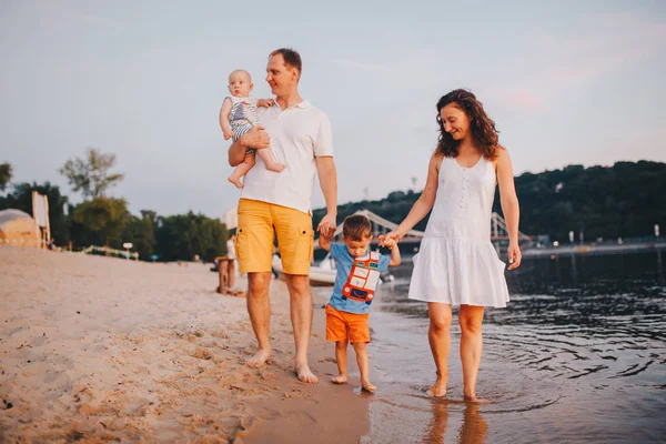 Vacaciones familiares en verano. Joven familia caucásica pie caminando descalzo playa de arena, ribera de agua del río. Papá mamá cogido de la mano dos hijos, hermanos. Gran familia amigable con dos niños cerca del lago —  Fotos de Stock