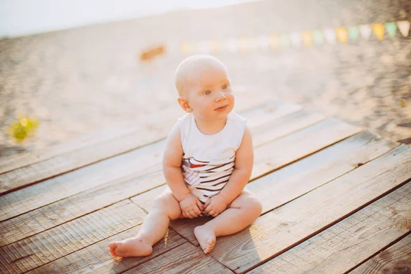 Niño un año rubio se sienta en un muelle de madera, un muelle con ropa a rayas, un complejo cerca del estanque en una playa de arena sobre un fondo de un río en el verano al atardecer del día —  Fotos de Stock