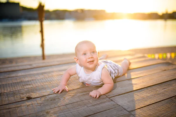 Un niño un niño, un año de edad, hombre rubio se acuesta sobre su estómago en el muelle de madera, muelle con ropa a rayas, compuesto cerca de estanque en la playa de arena contra el fondo del río en el verano al atardecer del día —  Fotos de Stock