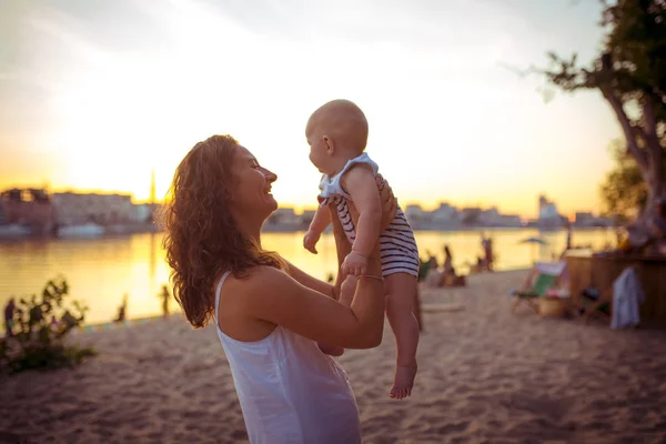 Joven mujer caucásica, hermosa madre sostiene a un niño en sus brazos, mamá abraza a su hijo de un año de edad, de pie en una playa de arena en un parque, cafetería al aire libre, cafetería de playa en el verano al atardecer —  Fotos de Stock