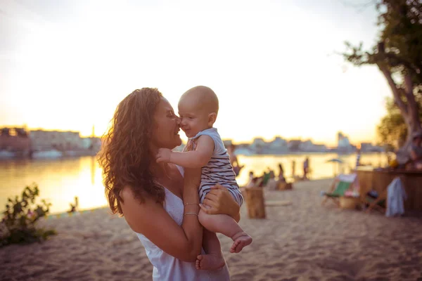 Joven mujer caucásica, hermosa madre sostiene a un niño en sus brazos, mamá abraza a su hijo de un año de edad, de pie en una playa de arena en un parque, cafetería al aire libre, cafetería de playa en el verano al atardecer —  Fotos de Stock