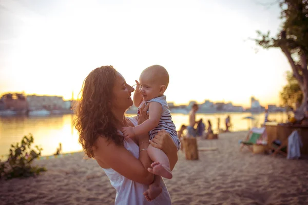 Joven mujer caucásica, hermosa madre sostiene a un niño en sus brazos, mamá abraza a su hijo de un año de edad, de pie en una playa de arena en un parque, cafetería al aire libre, cafetería de playa en el verano al atardecer —  Fotos de Stock