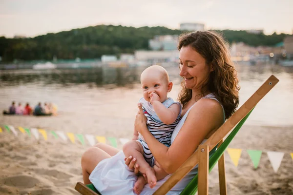 Joven mujer caucásica, hermosa madre sostiene al niño en sus brazos, mamá abraza a su hijo de un año sentado en una silla en la playa de arena en el parque, café al aire libre, café de playa en el verano al atardecer —  Fotos de Stock