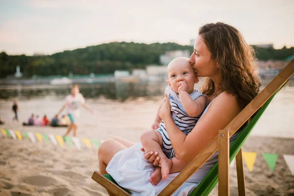 Joven mujer caucásica, hermosa madre sostiene al niño en sus brazos, mamá abraza a su hijo de un año sentado en una silla en la playa de arena en el parque, café al aire libre, café de playa en el verano al atardecer —  Fotos de Stock