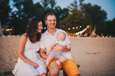 Sahilde aile yaz tatilleri teması. Beyaz adam ve kadın baba ve anne elinde oturan küçük bir bebek oğlu sarılmak. Genç aile gün batımında bir plaj kafede rahatlatıcı