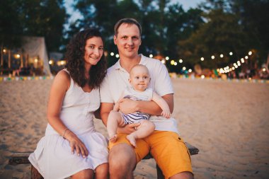 Sahilde aile yaz tatilleri teması. Beyaz adam ve kadın baba ve anne elinde oturan küçük bir bebek oğlu sarılmak. Genç aile gün batımında bir plaj kafede rahatlatıcı