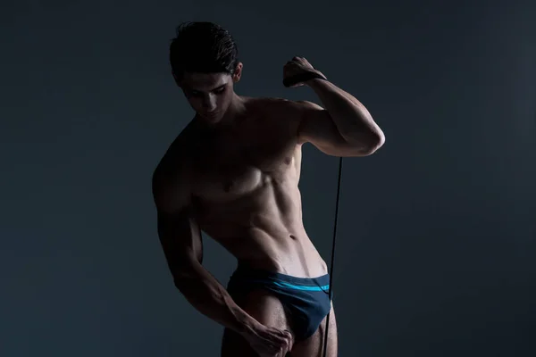 Muscular fitness young male antique perfect muscles six packs of abs and bare chest. Bodybuilder model trains with a stretching elastic against a dark background in the studio. Workout training gym