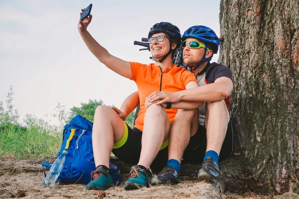 Щаслива пара з гірським велосипедом, що приймає смартфон селфі на відкритому повітрі. Щаслива пара, яка веде активний спосіб життя, зупинилася в лісі біля дерева, щоб сфотографуватися. Тематичний спорт та активний спосіб життя — стокове фото