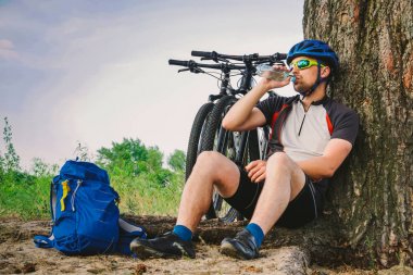 Beyaz erkek bisikletçi aktif yaşam tarzı şişe Bisiklet içme suyu egzersiz sonra ağaç altında istirahat. Kask ve spor sporcusu. El Flask. Doğaya susamış. Binicilik dağ bisikleti