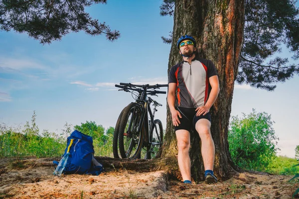 Кавказский велосипедист активный образ жизни отдыхает под деревом после занятий велосипедной питьевой водой из бутылки. Спортсмен в шлеме и спортивной одежде. Фляжка для рук. Жажда природы. Катание на горном велосипеде — стоковое фото