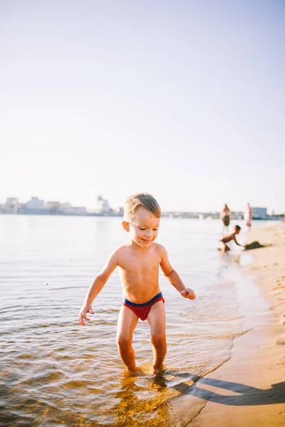 Летние развлечения на свежем воздухе у реки на городском пляже в Украине. Маленький смешной мальчик бежит вдоль берега реки, прыгая всплеск воды песчаный пляж летнее время — стоковое фото