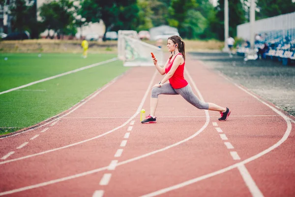 Sportovní fitness kavkazské ženy se zahřívat před cvičením. Sportovec natahoval nohy před tréninkem. Joggingu ve městě na stadionu. Dívka používá sluchátka s sluchátky telefon sportovní přísahy — Stock fotografie