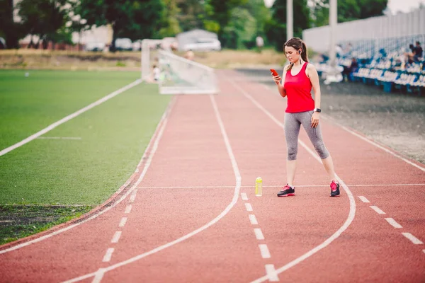 Sportovní fitness kavkazské ženy se zahřívat před cvičením. Sportovec natahoval nohy před tréninkem. Joggingu ve městě na stadionu. Dívka používá sluchátka s sluchátky telefon sportovní přísahy — Stock fotografie