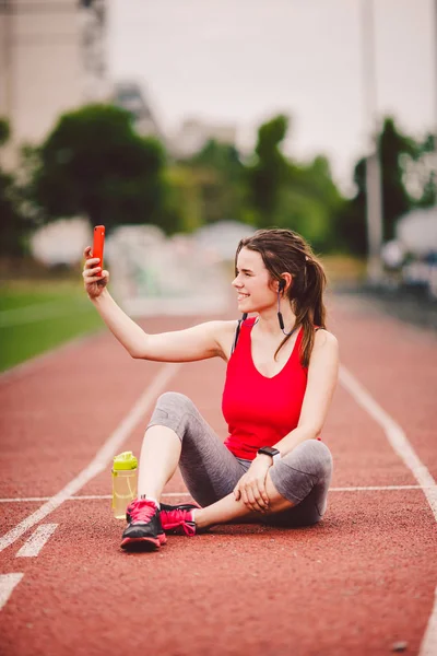 Mujer joven atleta en el estadio estilo de vida deportivo, sentada en la pista, tomando una foto selfie en un teléfono inteligente, sonriendo alegre en sus oídos auricular, cerca de un frasco de botella con agua — Foto de Stock