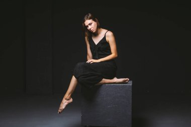Stüdyo siyah arka planda poz veren genç beyaz kadın model. Koyu renkli bir duvarda oturan siyah elbiseli kız. Denek kötü psikolojik durum, intra, sorunlar, kişilik çatışması