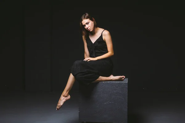 Young Caucasian female model posing in studio black background.Girl sitting in a black dress on a dark wall. Subject severe poor psychological state, intra, problems, personality conflict