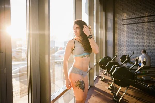 Mulher esportiva caucasiana jovem com cabelos longos encaracolados posa em sportswear perto de uma janela panorâmica em um clube de fitness. Jovem modelo atleta feminina com corpo bonito fica no ginásio com uma tatuagem — Fotografia de Stock