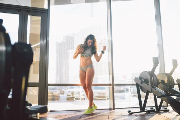 Joven mujer atlética caucásica con el pelo largo rizado posa en ropa deportiva cerca de una ventana del panorman en un club de fitness. Joven modelo atleta femenina con hermoso cuerpo se para en el gimnasio con un tatuaje —  Fotos de Stock