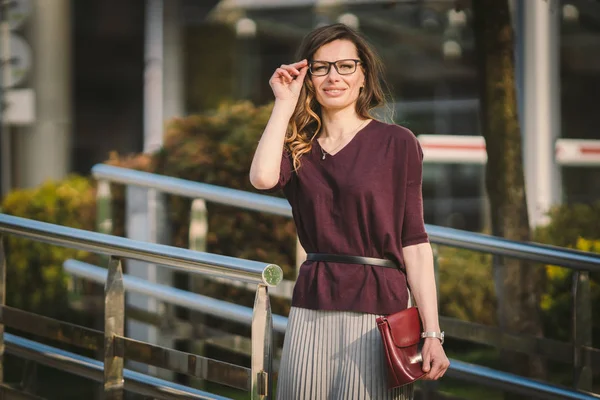 Caucasian adult lady woman in glasses posing near office building outside. Business lady in skirt holds hand glasses for vision bag in hand. Profession teacher assistant manager