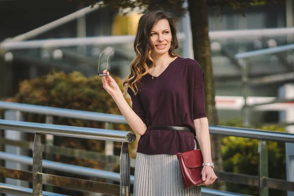 Mujer adulta caucásica en gafas posando cerca del edificio de oficinas afuera. Mujer de negocios en falda sostiene gafas de mano para la bolsa de visión en la mano. Profesor de profesión asistente gerente —  Fotos de Stock