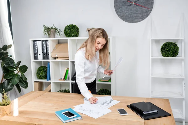 Mulher de negócios do tema no trabalho. Mulher branca bonita homem de negócios que trabalha de pé no escritório perto da mesa, verifica edições gráficos financeiros no papel — Fotografia de Stock