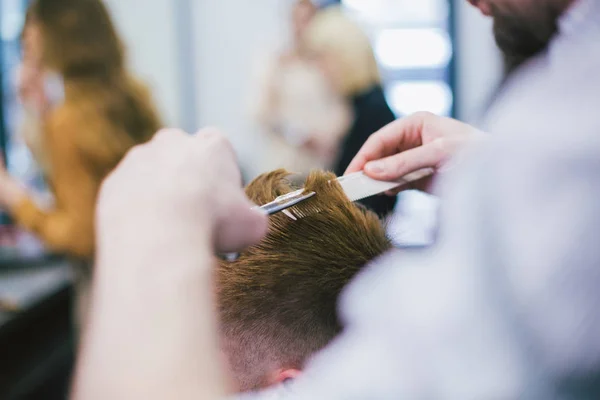 Парикмахер делает стрижку бородатого человека в парикмахерской. Профессиональный стилист стрижет волосы клиента в салоне. Парикмахер использует ножницы и расческу. Квалифицированный парикмахер стрижет мужчин. Концепция по уходу за волосами — стоковое фото