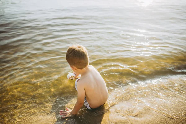 Petit garçon caucasien assis près d'une rivière sur une plage de sable et touche la surface de l'eau avec ses mains. Le thème est le flux du temps, une vie courte, le sens et le but de l'existence — Photo