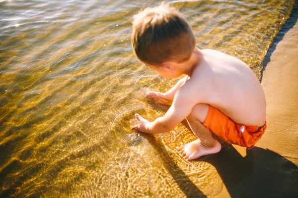 Petit garçon caucasien assis près d'une rivière sur une plage de sable et touche la surface de l'eau avec ses mains. Le thème est le flux du temps, une vie courte, le sens et le but de l'existence — Photo