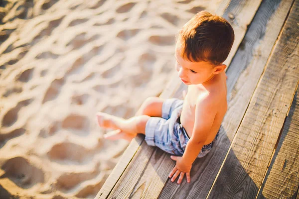 Маленький кавказский мальчик сидит на деревянном причале песчаного пляжа, летнее время, морской отдых у воды. Тема - течение времени, короткая жизнь, смысл и цель существования. Смысл жизни — стоковое фото