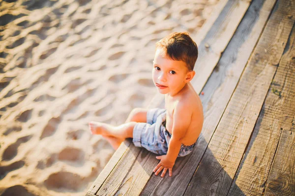 Little Caucasian boy child sitting on wooden pier sandy beach, summer time, sea vacation near water. The theme is the flow of time, a short life, the meaning and purpose of existence. Meaning of life