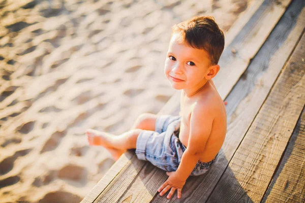 Little Caucasian boy child sitting on wooden pier sandy beach, summer time, sea vacation near water. The theme is the flow of time, a short life, the meaning and purpose of existence. Meaning of life