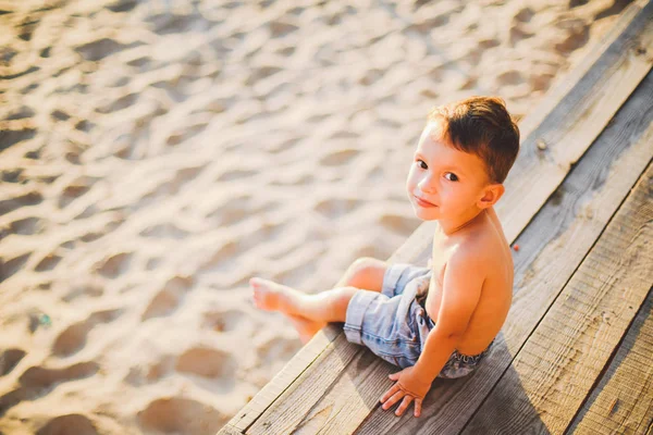 Niño pequeño caucásico sentado en la playa de arena del muelle de madera, hora de verano, vacaciones de mar cerca del agua. El tema es el flujo del tiempo, una vida corta, el significado y el propósito de la existencia. Significado de la vida —  Fotos de Stock