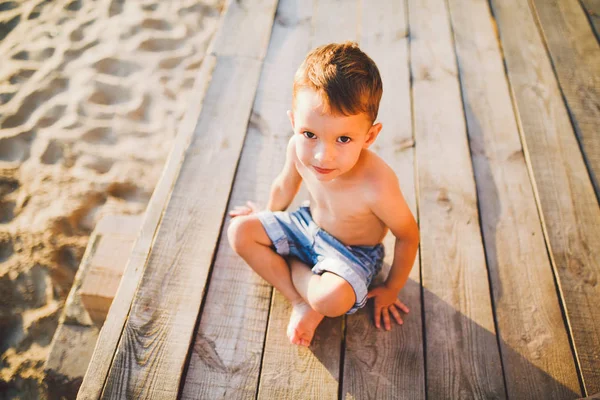 Niño pequeño caucásico sentado en la playa de arena del muelle de madera, hora de verano, vacaciones de mar cerca del agua. El tema es el flujo del tiempo, una vida corta, el significado y el propósito de la existencia. Significado de la vida —  Fotos de Stock