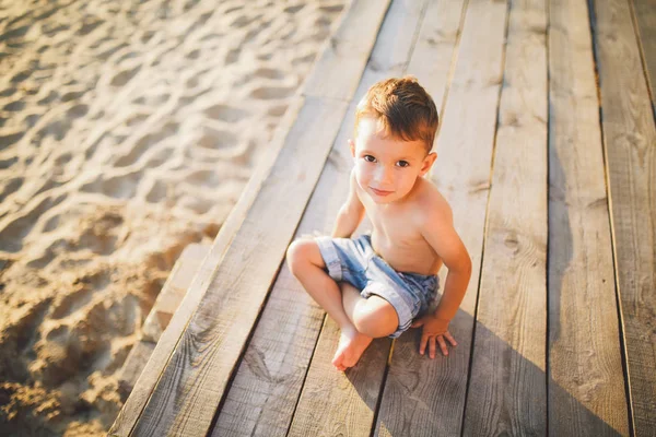 Petit garçon caucasien assis sur une jetée en bois plage de sable, heure d'été, vacances en mer près de l'eau. Le thème est le flux du temps, une vie courte, le sens et le but de l'existence. Signification de la vie — Photo
