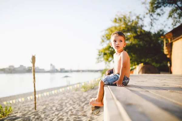 Niño pequeño caucásico sentado en la playa de arena del muelle de madera, hora de verano, vacaciones de mar cerca del agua. El tema es el flujo del tiempo, una vida corta, el significado y el propósito de la existencia. Significado de la vida —  Fotos de Stock