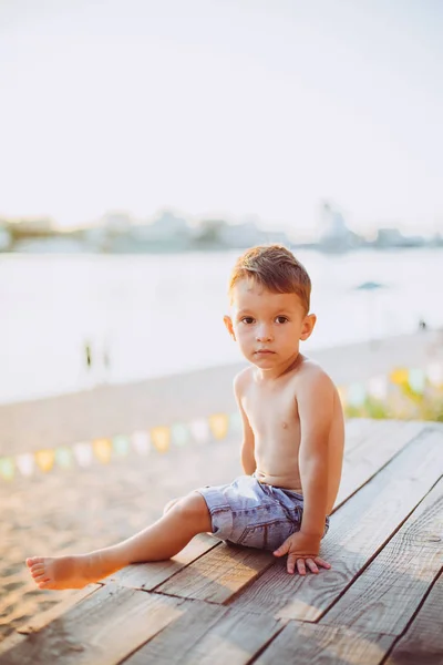 Niño pequeño caucásico sentado en la playa de arena del muelle de madera, hora de verano, vacaciones de mar cerca del agua. El tema es el flujo del tiempo, una vida corta, el significado y el propósito de la existencia. Significado de la vida —  Fotos de Stock