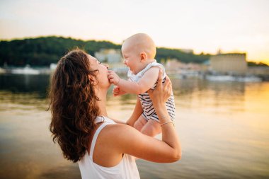 çocuğu gün batımına ve suya karşı döndüren anne. Mutlu anne ve bebek. Sahilde oynuyorum. Oğlunu havaya kaldıran genç kadın.